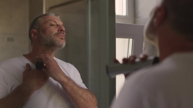 Handsome man shaving neck with electric trimmer standing near bathroom mirror