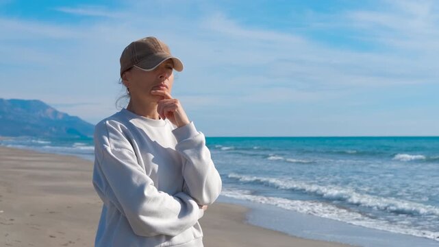 Pensive woman thinking alone on the beach. Thoughtful woman wearing a baseball cap and sweatshirt standing on a sandy beach and looking at the sea, contemplating life and feeling lonely
