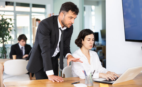 Male employee of the bank office stands near table and helps woman to fill out documents and bills on a laptop. Office employees are working on a project and checking data on a laptop