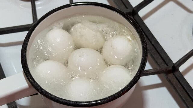 Close-up white eggs boiling in ladle on gas stove, seven eggs gently simmering in enamel ladle with blue flame beneath, steam