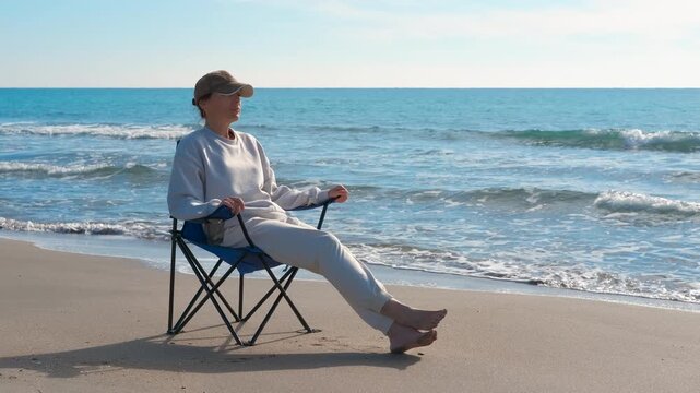 Woman relaxing on a beach chair watching the ocean waves. Solitary woman sitting in a foldable chair on a sandy beach, relaxing barefoot while enjoying the serene view of the sea waves