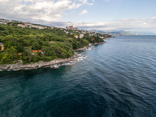 Fototapeta premium Rijeka, Croatia - 05 18 2025: Angled drone photo showing the shore of Gradska plaža Razbojna beach on the left, with wavy Adriatic Sea hitting the rocky shore on the right, buildings and mountains
