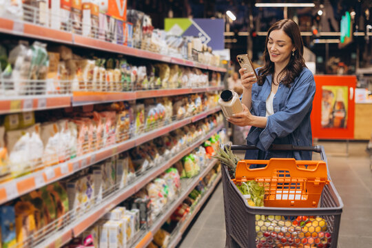 Woman in supermarket aisle checking product information on smartphone while grocery shopping
