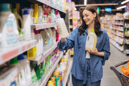 Woman standing in a grocery store, holding and comparing two bottles of liquid detergent while shopping for household items