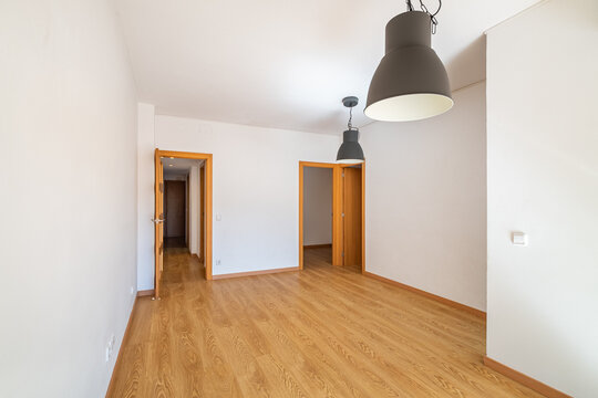 Empty room with wooden floor, white walls, and two pendant lights. Doors lead to other spaces. Bright and spacious atmosphere.