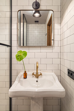 A modern bathroom with a white sink, gold faucet, and a green plant in a brown vase. The walls are tiled with white subway tiles and a mirror is mounted above the sink.