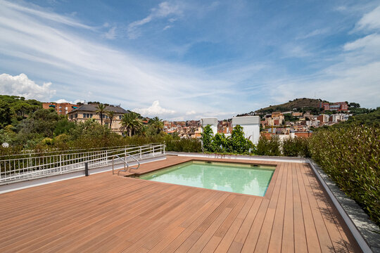 A modern rooftop terrace with a swimming pool. The area features wooden decking and overlooks a scenic view of buildings and hills under a clear blue sky.