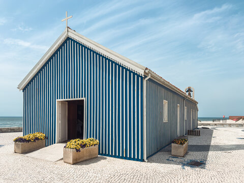 Perspective view of the iconic blue and white striped chapel of Praia de Mira nestled against the Atlantic ocean under a beautiful clear blue sky