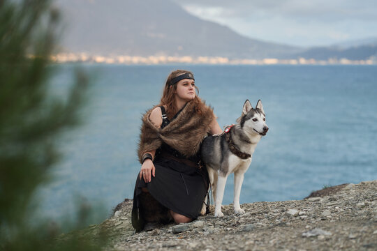 A woman sits beside a Siberian Husky dog on coastal rocks. Blue sea mountain landscape and relaxed posture create peaceful travel lifestyle atmosphere outdoors.