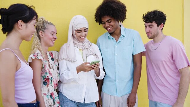 Group of young multicultural friends smiling and laughing while watching something on a smartphone against a vibrant yellow background