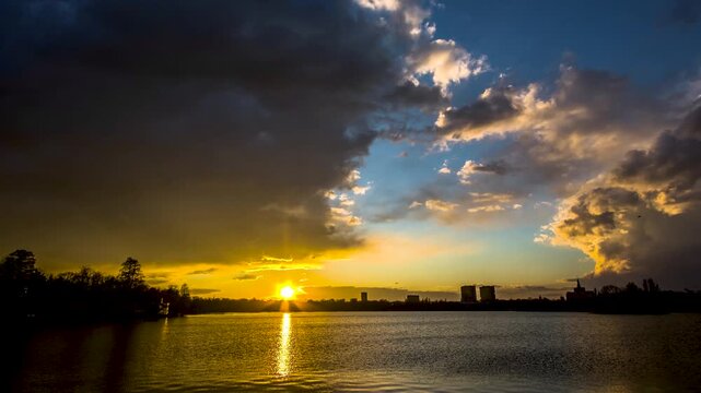 Bucharest, Romania - 01 October 2020: View of sunset over a lake with golden light reflecting on the water and a city skyline under dramatic clouds.