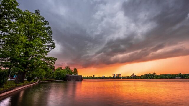Bucharest, Romania - 22 May 2020: View of dramatic storm clouds over Herastrau Lake at sunset with lush green trees and city skyline in the distance.