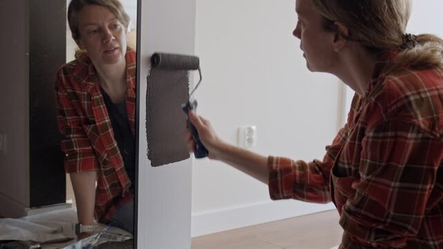 Woman applies dark paint to doorway with small roller beside mirror and protective sheet. Concept of DIY renovation and room decorating