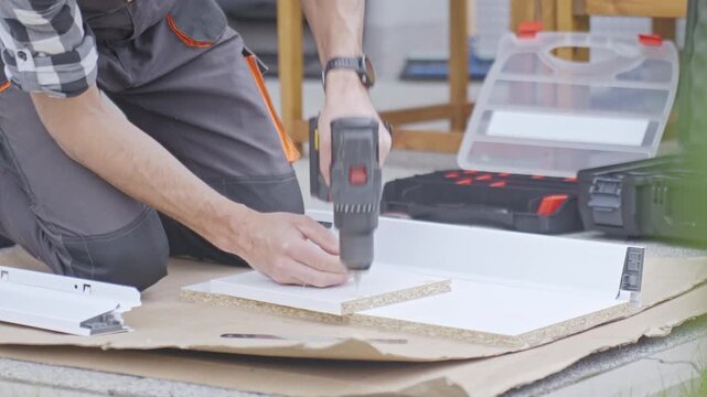 Man drills holes in drawer panel with cordless drill on cardboard covered floor. Concept of DIY furniture assembly and repair