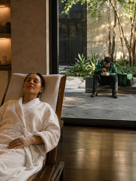 Woman relaxing in a spa robe indoors while a delivery worker eats outside the window, visual contrast of comfort, service labor, and urban inequality.