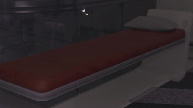 Secure infirmary bench under harsh light, worn red mattress with reinforced rails, visible restraint straps and concrete surroundings, sparse medical equipment