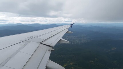 Obraz premium Airplane wing with flaps extended, soaring above green mountains and overcast sky during flight