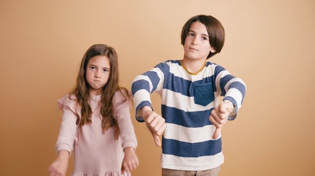 Young boy and a girl expressing disapproval and negativity with thumbs down gestures. Unhappy siblings looking at the camera with dislike