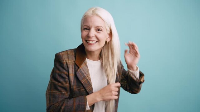 Happy mature woman with long platinum blonde hair posing, dancing, and smiling at the camera against a plain turquoise background