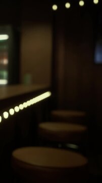 A bar scene shows empty stools lined up near a countertop. Soft light from the bar highlights the area. A vending machine is visible in the background. The setting feels intimate and inviting.