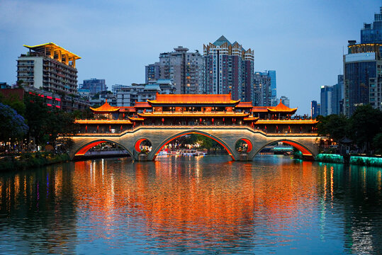 Symmetrical night view of the illuminated traditional Anshun covered bridge over Jin River with skyscrapers background, Chengdu, Sichuan, China