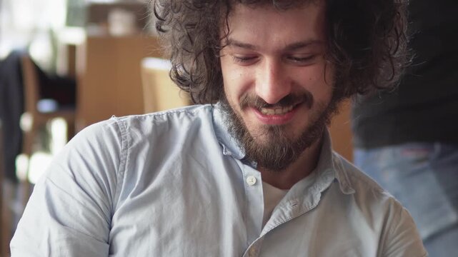 Cheerful man with curly hair and beard smiles warmly in a casual indoor environment holding smartphone