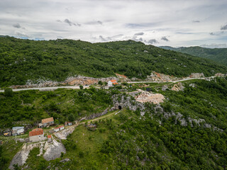 Fototapeta premium Slansko Lake, Montenegro -05 29 2025: Drone landscape photo of hilly terrain in Ponikvica, showing winding road, rural houses with red roofs, rocky outcrops, tunnel entrance, and a quarry under clouds
