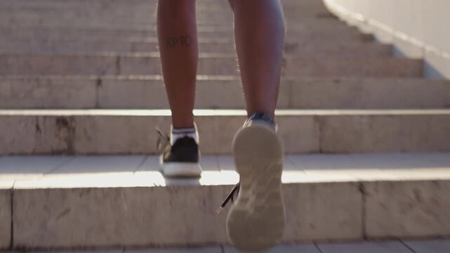 Woman in sportswear running up staircase during sunny outdoor workout. Back view of athletic female jogger training on stairs with strong sunlight beam
