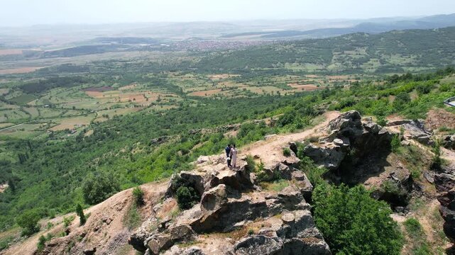 Newlywed romantic couple standing on scenic mountain summit cliff overlooking vast verdant landscape