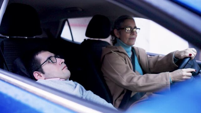 Mature woman driving a car with a young man having a physical condition in the passenger seat. Concept of daily life, support, and mobility