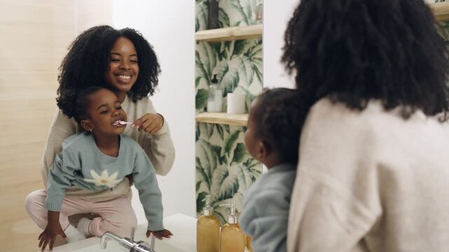 Young mother teaching her cute little daughter about oral hygiene. Happy female helping a child to brush her teeth in the bathroom mirror