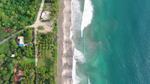 Stunning aerial top down view of turquoise ocean waves gently lapping a sandy beach next to a lush tropical jungle with palm trees in costa rica