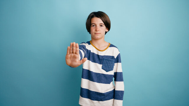 Boy making an open palm stop gesture, showing refusal and warning. Standing against a blue background, expressing boundaries