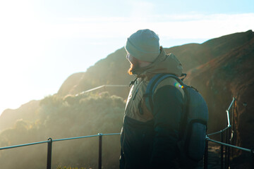 A male hiker with a backpack stands on a stone mountain path, looking out over a misty, sunlit alpine landscape. © Maria Sap