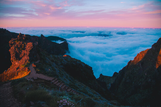 A scenic stone hiking trail splits on a mountain ridge high above a dense sea of clouds at pink sunset.