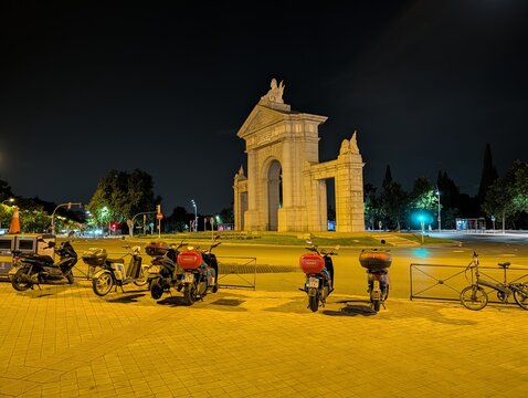 25 May 2026. Madrid, Spain. Puerta de San Vicente monument illuminated at night in the Principe Pio area of Madrid, with several shared rental motorcycles parked along the sidewalk in the foreground.