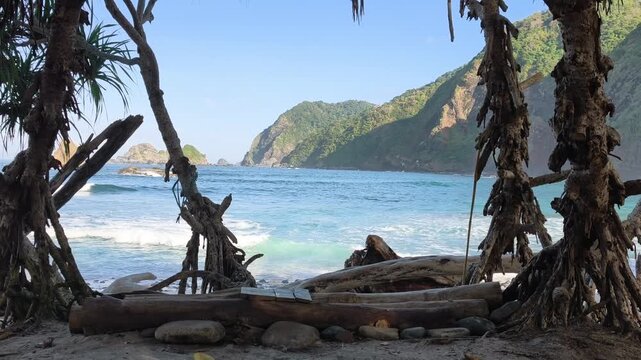 Scenic coastal view framed by tree branches with natural wave sound. Blue ocean waves crashing against the shore with rocky hills and islands under a sunny sky. Papuma Beach, Indonesia.