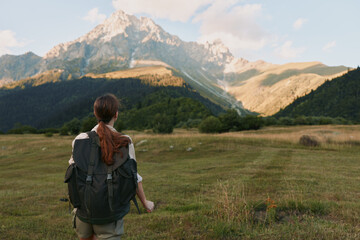 Fototapeta premium Woman hiker with backpack stands in a grassy valley, mountains ahead under a sunlit sky, conveying adventure and calm in a natural outdoor setting