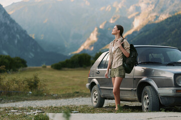 Fototapeta premium Young woman with backpack enjoying a mountain landscape beside a parked car, gazing into the distance. A carefree outdoor lifestyle captures adventure, travel, and nature in warm, soft light on