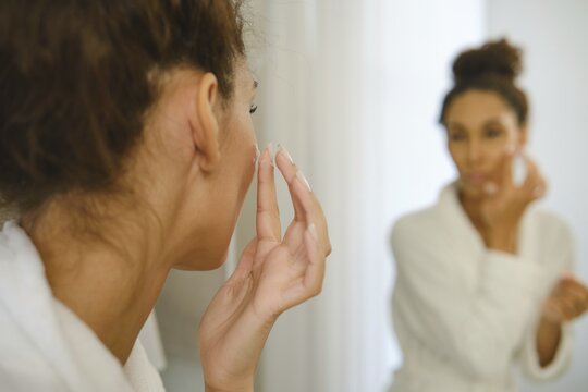 A woman in a white robe is applying cream to her face while looking at herself in the bathroom mirror. Soft lighting creates a serene atmosphere as she follows her morning skincare regimen