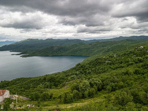 Slansko Lake, Montenegro - 05 29 2025: Drone photo of Slansko Lake/Slano Jezero with deep blue water, green hills, rustic stone house, and irregular shoreline under a cloudy sky.
