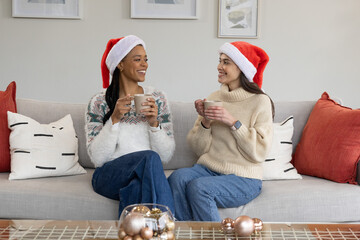 Fototapeta premium African American and Asian women chatting on gray sofa wearing Santa hats, holding ceramic mugs