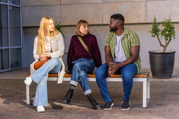 Obraz premium Friends sitting on wooden bench by tiled wall, chatting while holding brown handbag and green bag