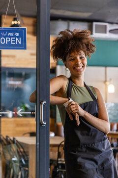 African American adult woman smiling and adjusting watch in cafe doorway with black apron OPEN sign