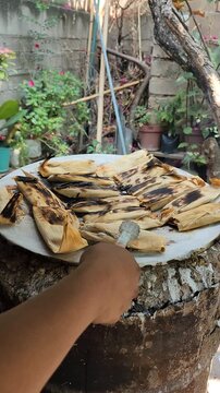 Worker carefully turning over toasted tamales with charred corn husks on a clay comal