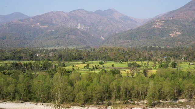 View of a riverbank with lush green trees and agricultural fields against a backdrop of rolling mountains in Nepal.