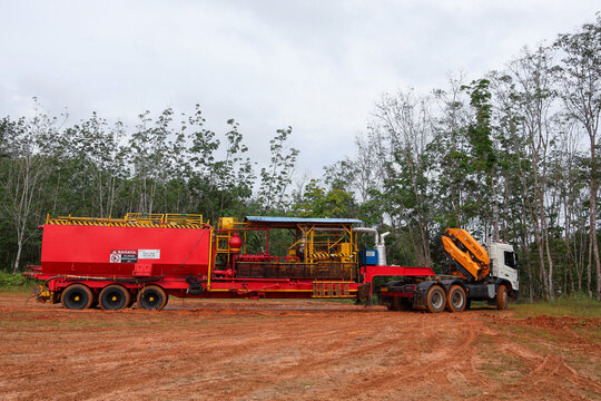 ​A large red oilfield utility service trailer equipped with a fluid tank unit attached to a white truck with a hydraulic crane arm at a remote petroleum drilling site.