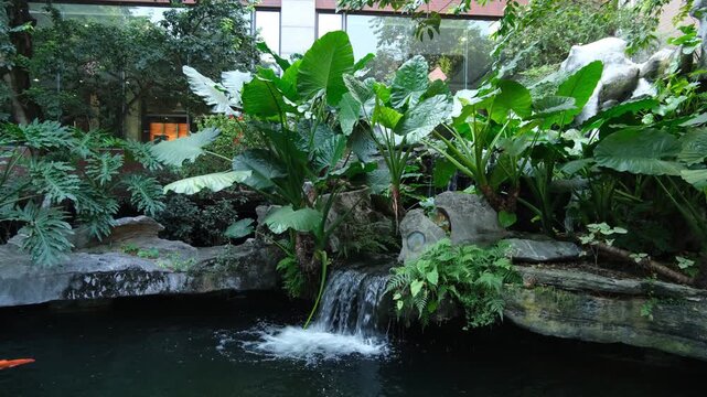 A landscaped indoor atrium or courtyard garden featuring a tiered artificial rock waterfall cascading into a koi pond, lush tropical foliage, decorative rock formations