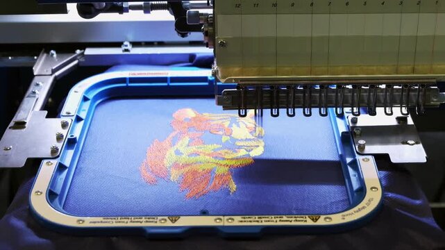 Industrial embroidery machine creating colorful tiger face on blue textile material with precision.