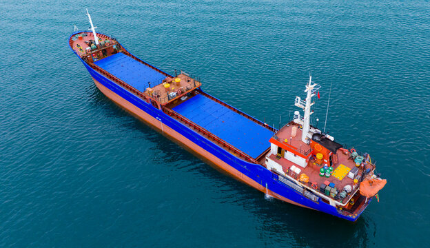 High-angle aerial drone shot of a massive merchant cargo vessel with distinctive blue hatch covers cruising on dark ocean water. Showcasing international freight and trade industry.